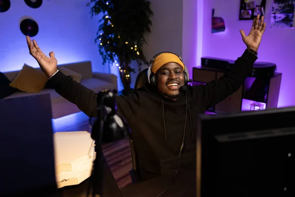 Mutlu oyuncu, elinde gülümsemeyle video oyunu kazanmaktan zevk alıyor. Günlük kıyafetler ve kulaklık takıyor. Afiş kazananı, renkli ışıkları olan bilgisayar karanlık arka planında evde oynar