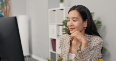 Kareli ceket giyen, uzun kahverengi saçlı odaklanmış güzel bir kadın bilgisayar ekranının önünde masasında oturuyor, haberleri, makaleleri, belgeleri okuyor, çene altı destekleyen elleri analiz ediyor, gülümsüyor.