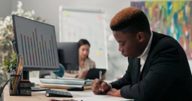 Takım elbiseli koyu tenli bir adam bilgisayar monitörünün önündeki masada oturur, kağıda basılmış grafikleri tanımlar, onları ekrandaki diyagramlarla karşılaştırır, imzalar, uyumluluklarını onaylar..