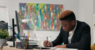 Takım elbiseli koyu tenli bir adam bilgisayar monitörünün önündeki masada oturur, kağıda basılmış grafikleri tanımlar, onları ekrandaki diyagramlarla karşılaştırır, imzalar, uyumluluklarını onaylar..