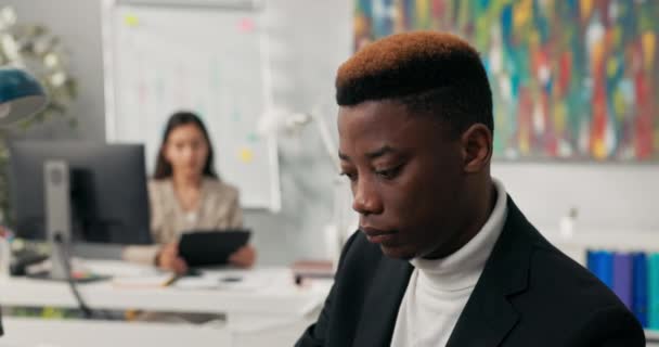 Homem de pele escura em jaqueta com cabelos coloridos trabalha no escritório da empresa preenche documentos sobre computação sem discriminação em segundo plano mulher está respondendo e-mail em tablet, trabalho diário na empresa. — Vídeo de Stock