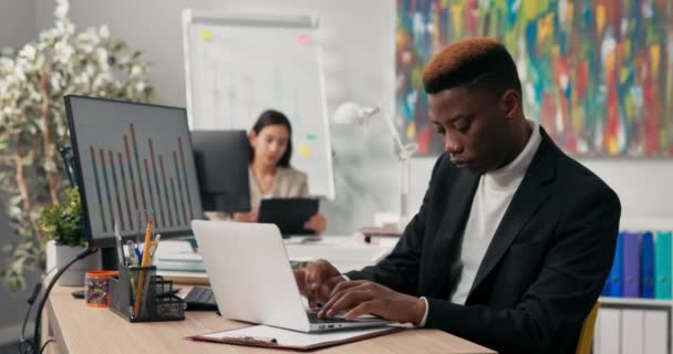 A smiling dark-skinned boy spends morning at work in front of computer screen, tapping fingers on laptop keyboard, filling out official and company applications, comparing correctness on two monitors. — Stock Video