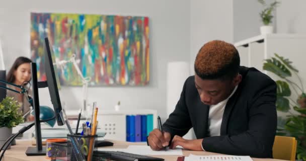 黑皮黑衣男子坐在电脑监控器前的办公桌前，描述印在纸上的图表，并与屏幕上的图表相比较，指出，背景中的同事. — 图库视频影像