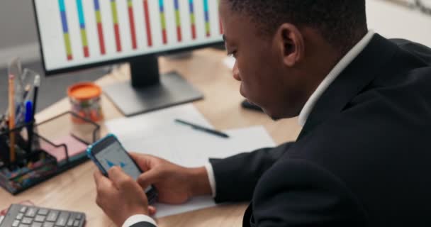 Un hombre de piel oscura vestido con un traje se sienta en un escritorio frente a un monitor de computadora, sosteniendo un teléfono en las manos, revisando gráficos de barras, gráficos, trabajando en estadísticas de la compañía, oficina de análisis. — Vídeos de Stock
