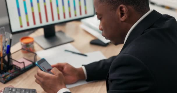 Un hombre de piel oscura vestido con un traje se sienta en un escritorio frente a un monitor de computadora, sosteniendo un teléfono en las manos, revisando gráficos de barras, gráficos, trabajando en estadísticas de la compañía, oficina de análisis. — Vídeos de Stock