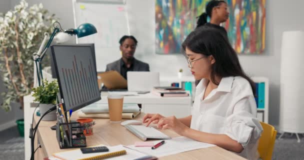 Hermosa secretaria en gafas con característica belleza asiática coreana vestida con camisa blanca, se sienta en la oficina frente a la computadora, trabajando, en el fondo compañeros de trabajo caminan alrededor de la empresa. — Vídeo de stock