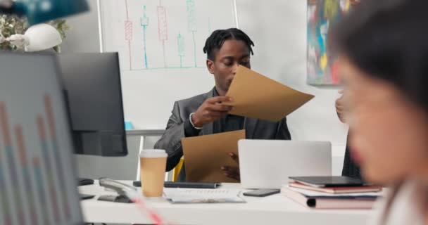 スーツを着た忙しい暗い肌のボスは、コンピュータの机の後ろに座って、アシスタントは、彼の封筒、手紙、男が開き、対応を通して見て、女性に戻り、机の上の葉を与えます. — ストック動画