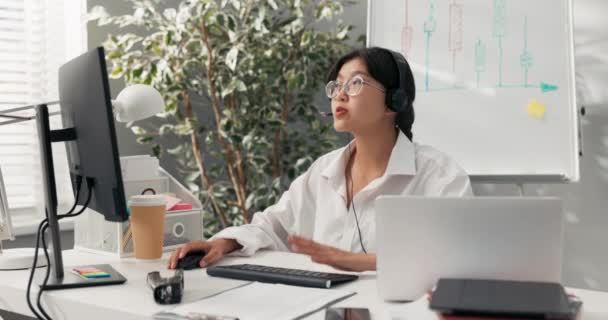 Krásná elegantní žena v brýlích s asijské korejské krásy sedí v kanceláři společnosti s sluchátky mikrofon na uších před počítačem vede konzultace s klientem pojištění vysvětluje.