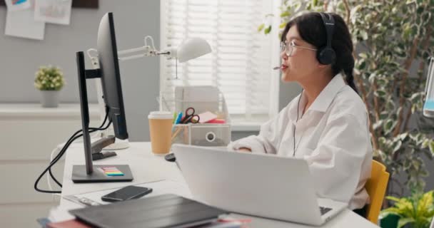Całkiem eleganckie kobiety w okularach z azjatycką koreańską urodą siedzi w biurze firmy ze słuchawkami mikrofon na uszach przed komputerem prowadzi konsultacje klienta ubezpieczenie wyjaśnia uśmiech. — Wideo stockowe