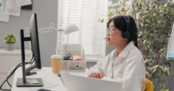 Elegantní ženy v brýlích s asijskou korejskou krásou sedí v kanceláři společnosti s sluchátky mikrofon na uších před počítačem vede konzultace s klienty pojištění vysvětluje úsměv. — Stock video