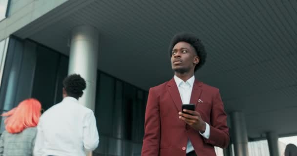 Chefen för företaget, en mörkhyad man i kostym som arbetar på ett kontor, går ut ur en modern företagsbyggnad, håller en telefon i handen, kollar nyheterna, tittar på ny information på Internet. — Stockvideo