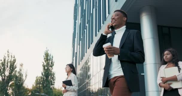 Συνάδελφοι της εταιρείας σπάνε ο ένας τον άλλο έξω από την είσοδο του μοντέρνου κτιρίου σκουρόχρωμος άντρας ντυμένος με σακάκι βγαίνει από το γραφείο μιλώντας στο τηλέφωνο κρατώντας ένα φλιτζάνι καφέ μιας χρήσης.. — Αρχείο Βίντεο