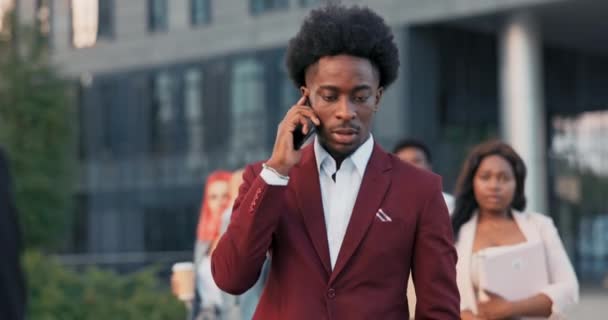 Красавчик уверенный в себе афроамериканский бизнесмен топ-менеджер в стильном коричневом костюме, выходит на перерыв на переговоры по телефону на фоне бизнес-центра, офиса компании — стоковое видео