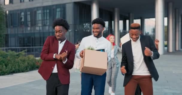 Co-workers change workstations, three dark-skinned men at front dance happy, middle one carries a box with their stuff, girls following behind are also funny, playing, waving their hands in the air — Stock Video