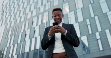 Ceket giymiş, gülümseyen mutlu çocuk camdan yapılmış modern şirket binasının önünde duruyor, işten çıkıyor, telefonu tutuyor, mesajlar yazıyor, mesajlar yolluyor, tebligatları okuyor.