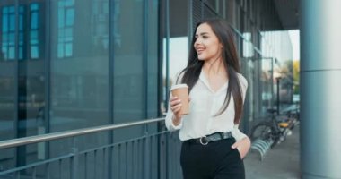 Güzel, neşeli bir kadın bir ofiste çalışıyor, şirket, şirket, zarif giysiler içinde cam bir binanın önünde yürüyor, etrafa bakıp gülümsüyor, kahve molası veriyor, görevinden uzaklaşıyor.