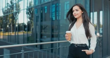 Güzel, neşeli bir kadın bir ofiste çalışıyor, şirket, şirket, zarif giysiler içinde cam bir binanın önünde yürüyor, etrafa bakıp gülümsüyor, kahve molası veriyor, görevinden uzaklaşıyor.