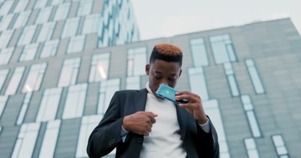Темношкірий чоловік, одягнений у костюм, стоїть перед скляною будівлею компанії, співробітник закінчує дзвінок по телефону з абонентом ставить смартфон у кишенькові клаптики руки танці щаслива добра інформація — стокове відео