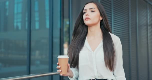 Ragazza attraente che lavora in un ufficio, società, società, vestita con abiti eleganti cammina di fronte a un edificio di vetro, si guarda intorno, pausa caffè, riposo dai suoi doveri — Video Stock