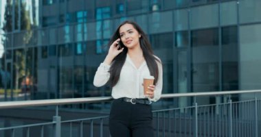 Zarif bir şekilde giyinmiş çekici bir kadın bir şirkette çalışıyor, bir şirket, cam modern bir binanın önünde yürüyor, telefonla konuşuyor, elinde bir fincan kahveyle gidiyor.