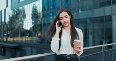 Zarif bir şekilde giyinmiş çekici bir kadın bir şirkette çalışıyor, bir şirket, cam modern bir binanın önünde yürüyor, telefonla konuşuyor, elinde bir fincan kahveyle gidiyor.