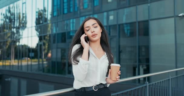 Een mooi meisje leunt tegen een leuning voor een modern glazen kantoorgebouw, een zakenvrouw gekleed in een wit shirt houdt een telefoon aan het oor, praat met een vriend — Stockvideo