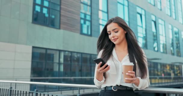 Güzel bir iş kadını modern cam şirket binasının parmaklıklarına yaslanmış duruyor, bir kız elinde bir telefon tutuyor, mesajlar yazıyor, kahve fincanından bir yudum alıyor. — Stok video