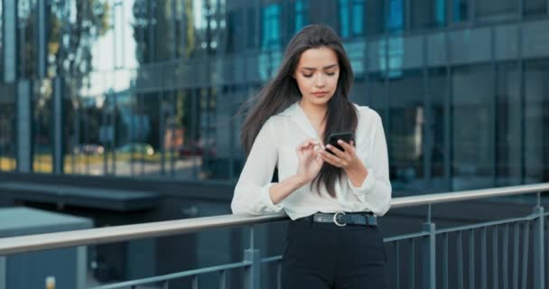 Mooie glimlachende vrouw werkt in gezelschap komt uit voor pauze gekleed in elegante outfit staat leunend tegen leuning in de voorkant van glazen gebouw met telefoon in de hand leest nieuws barst lachen — Stockvideo