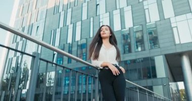 Beyaz gömlekli güzel çekici bir kadın çalıştığı şirketin cam binasının yanında duruyor. İş kadını önüne bakıyor, saçları dalgalı, gülümsüyor.