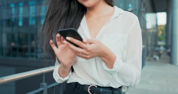 Close-up shot of neat hands of elegant woman holding phone, smartphone, woman taps screen, sends message, dressed in shirt walks past glass building of company, business affairs — Stock Video