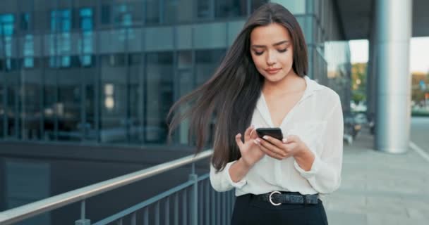 Beautiful attractive elegant woman holding a phone in her hands, smartphone, girl taps on the screen, sends a message, wearing a shirt walks past glass building of the company, business affairs — Stock Video