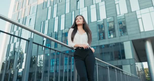 Hermosa atractiva elegantemente vestida en camisa blanca mujer se para en la barandilla junto al edificio de vidrio de la corporación donde trabaja, mujer de negocios mira hacia el futuro, cabello desaliñado, sonriendo a la cámara — Vídeos de Stock