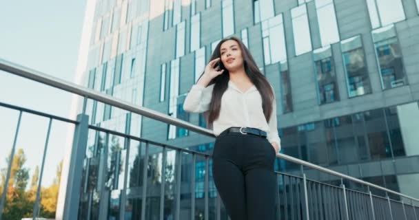 Красивая девушка стоит, прислонившись к перилам перед современным стеклянным офисным зданием, деловая женщина в белой рубашке держит телефон к уху, разговаривает с другом, улыбается — стоковое видео