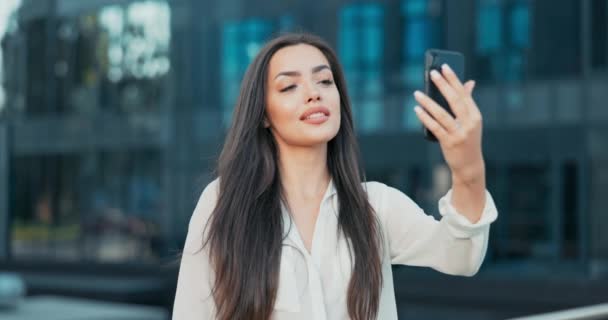 Uma menina bonita fica encostada a um corrimão na frente de um prédio de escritórios de vidro moderno, uma mulher de negócios vestida com uma camisa branca segura um telefone na frente de conversas através de uma câmera de vídeo com um amigo — Vídeo de Stock