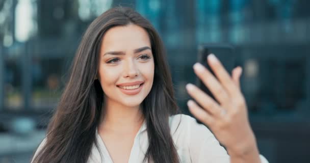 Una bella ragazza si trova appoggiata ad una ringhiera di fronte ad un moderno edificio per uffici in vetro, una donna d'affari vestita con una camicia bianca tiene un telefono davanti ai colloqui attraverso una videocamera con un amico — Video Stock