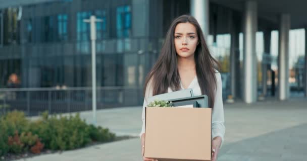 Žena kráčí před skleněnou moderní kancelářskou budovou korporace, kde pracovala, byla propuštěna ze své pozice dívka se smutným nejistým výrazem na tváři drží krabici sbalených věcí nezaměstnanost — Stock video