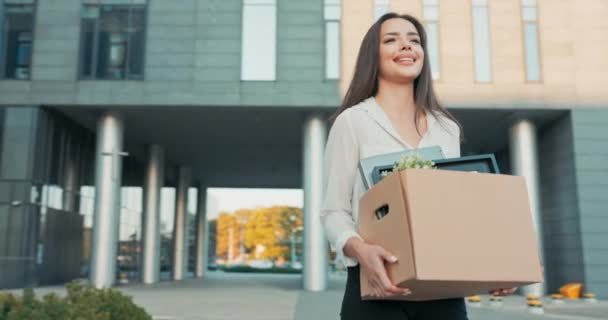 Atraktivní krásná usměvavá bruneta v košili stojí před skleněnou moderní firemní budovou v rukou drží krabici zabalených věcí, povýšení na pozici, přichází do nové práce v kanceláři — Stock video