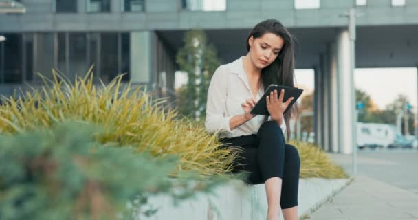 Seorang wanita tersenyum, karyawan perusahaan, sekretaris, duduk istirahat di depan gedung perusahaan, dinding tanaman, seorang wanita berambut cokelat memegang tablet di tangan, browsing media sosial, surfing internet — Stok Video