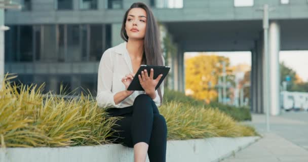 Mujer bonita, empleada de la empresa, secretaria, se sienta en el descanso frente a un edificio corporativo, una pared de plantas, una mujer morena sostiene una tableta en las manos, navegar por las redes sociales, navegar por Internet — Vídeo de stock