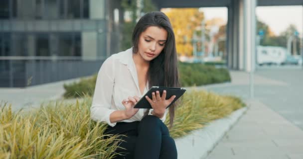 Donna, dipendente dell'azienda, segretaria, si siede in pausa di fronte a un edificio aziendale, un muro di piante, una donna bruna tiene un tablet in mano, navigando sui social media, navigando su Internet — Video Stock