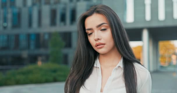 Atractiva hermosa morena fotogénica con ojos oscuros en maquillaje diario mira a la cámara, mujer seductora, vestida con una camisa elegante, mujer de negocios de pie frente a un edificio corporativo — Vídeos de Stock