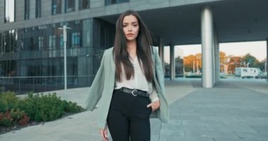 Kahverengi saçlı, sırıtan bir kız, şık gömlek ve ceket giymiş, camdan gökdelenin, sekreterin, iş kadınının, patronunun önünden geçerken, mola vermek için şirket binasından çıkıyor.