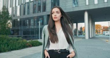 Kahverengi saçlı, sırıtan bir kız, şık gömlek ve ceket giymiş, camdan gökdelenin, sekreterin, iş kadınının, patronunun önünden geçerken, mola vermek için şirket binasından çıkıyor.