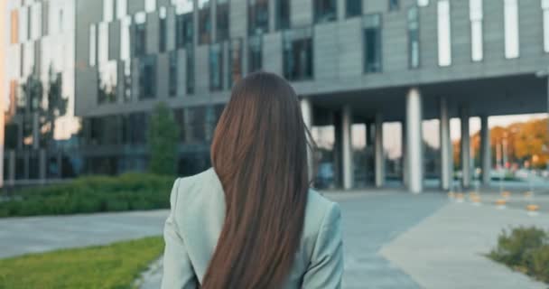 Disparo por detrás de una mujer elegante, morena de pelo largo, chica regresa de la rotura al edificio de oficinas de cristal, se gira detrás de ella, mira a la cámara, continúa adelante — Vídeos de Stock