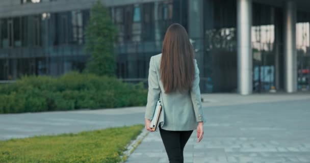 Disparo por detrás de la mujer, morena con el pelo largo, chica regresa de la rotura al edificio de oficinas de vidrio gira detrás de sus miradas en la cámara sigue caminando por delante carpetas de archivos de marcha sexy con documentos en la mano — Vídeos de Stock