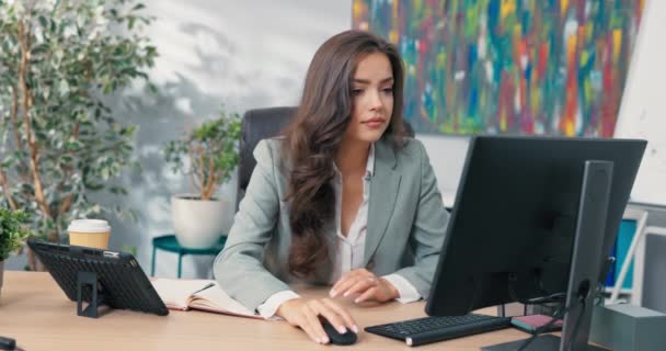 Focado no trabalho menina atraente em maquiagem clássica vestindo uma camisa e casaco, senta-se em uma mesa em uma empresa na frente de um computador, preenche documentos, lê e-mail, responde mensagens, sorrindo para a câmera — Vídeo de Stock