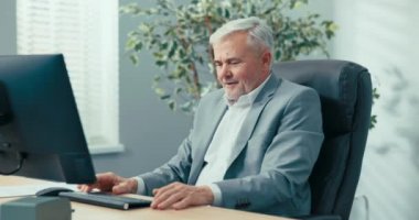 Gri saçlı zarif adam ofisten uzaktan konferans düzenliyor, dizüstü bilgisayarın önünde oturuyor, meslektaşlarına el sallıyor, web kamerasında online iş görüşmeleri yapıyor, görüntülü sohbet yapıyor.