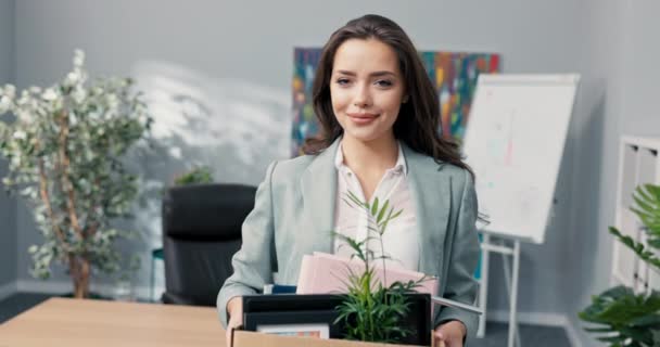 Привлекательная красивая улыбающаяся брюнетка в рубашке стоит перед столом в офисе компании в руках держит коробку с упакованными вещами, продвижение по службе, приход на новую работу в офис, трудоустройство — стоковое видео