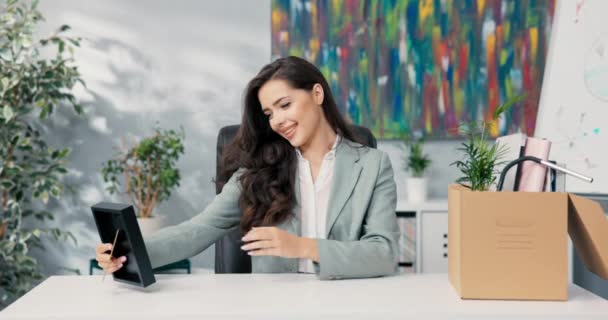 Karyawan baru di perusahaan, duduk di meja kantor, seorang sekretaris cantik membongkar kotak kardus peralatan, mengambil aksesoris di atas meja, set frame, tanaman, komputer terbuka — Stok Video