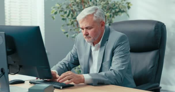 Reifer, grauhaariger, eleganter Geschäftsmann sitzt im Büro am Computer, vervollständigt Firmendokumente, überprüft Daten im System, sortiert Rechnungen, übernimmt Führungsaufgaben — Stockvideo